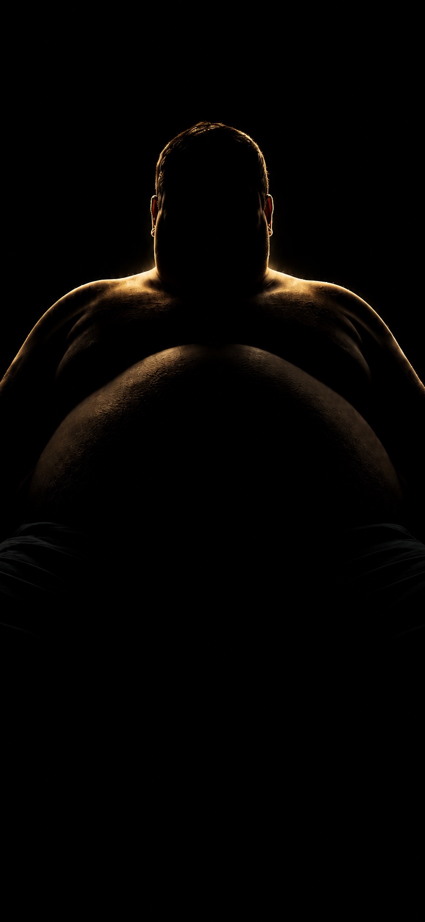 A large man seated in shadow, rim-lit from behind against a black background.